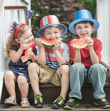 three children eating watermelon wearing patriotic gear