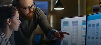 Two people staring at computer code on a dual screen monitor