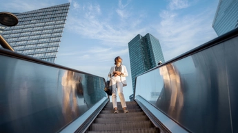 Woman on escalator going down with buildings and sky behind her