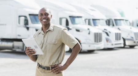A man standing in front of a row of parked semi-trucks outside a distribution warehouse, holding a digital tablet.